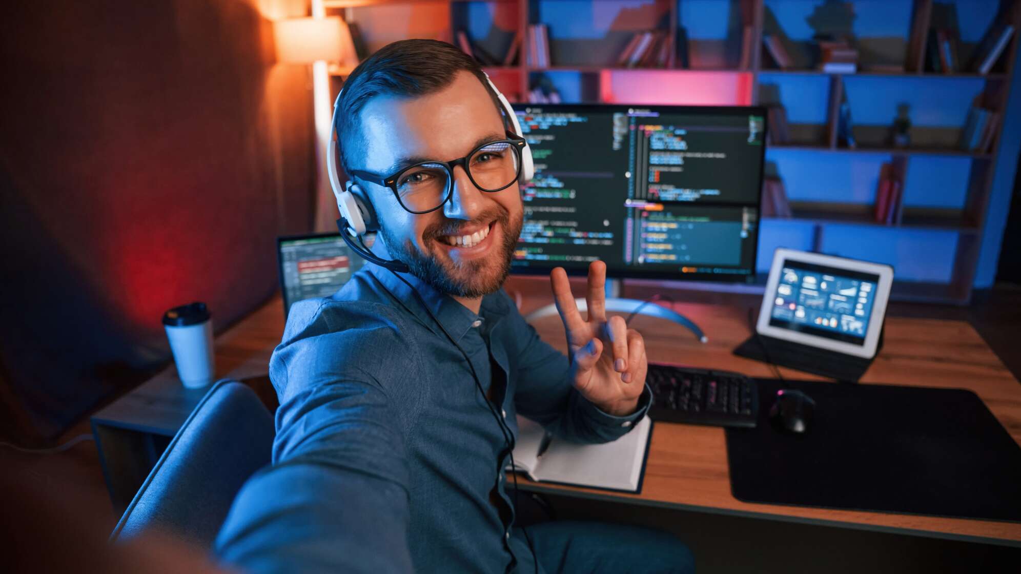 Ein Programmierer sitzt mit Headset an einem Schreibtisch vor mehreren Monitoren mit geöffnetem Code und zeigt ein Peace-Zeichen. Im Hintergrund sind Bücherregale und eine warme Beleuchtung zu sehen.